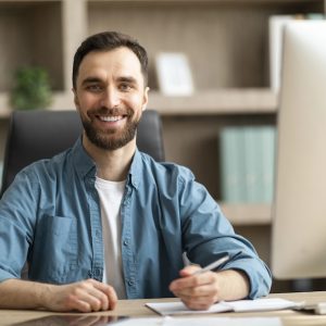 Business Person. Portrait Of Handsome Young Businessman Posing At Workplace In Office, Millennial Male Entrepreneur Sitting At Desk And Taking Notes, Looking And Smiling At Camera, Copy Space